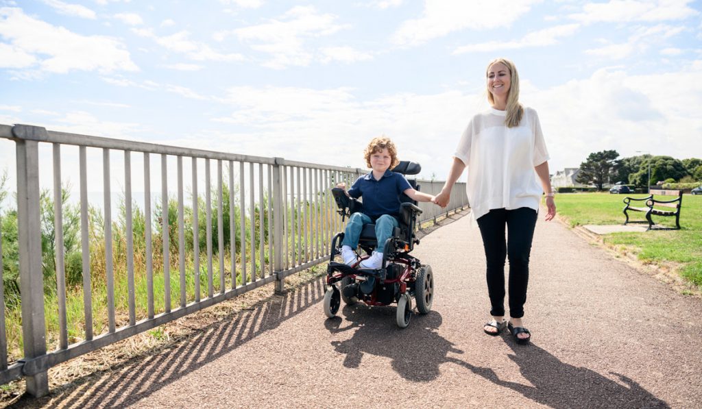Woman walks alongside a young boy riding an electric wheelchair.