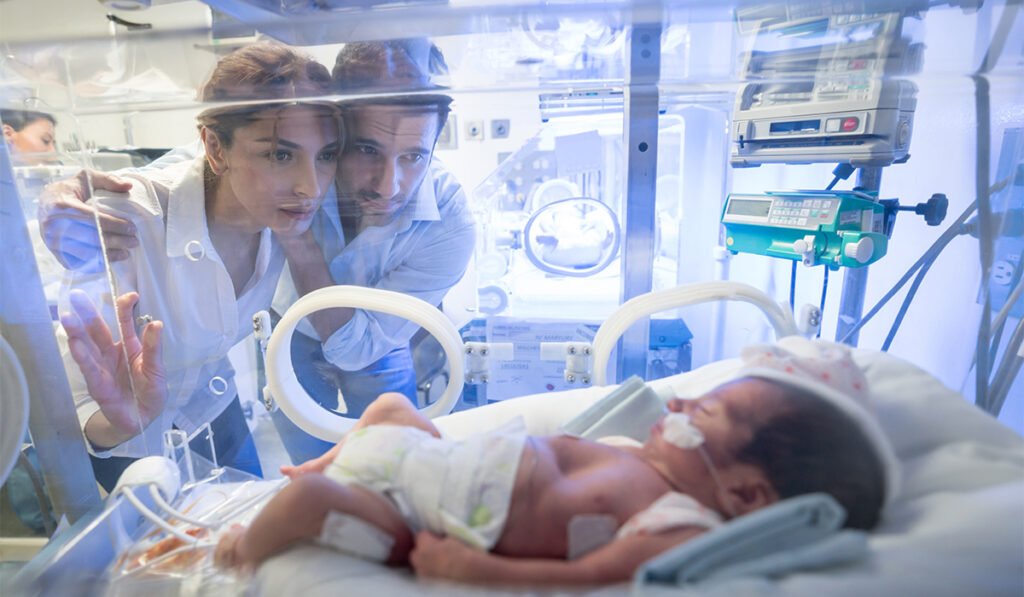 Worried young couple looking at their premature newborn in an incubator with oxygen at neonatal intensive care unit