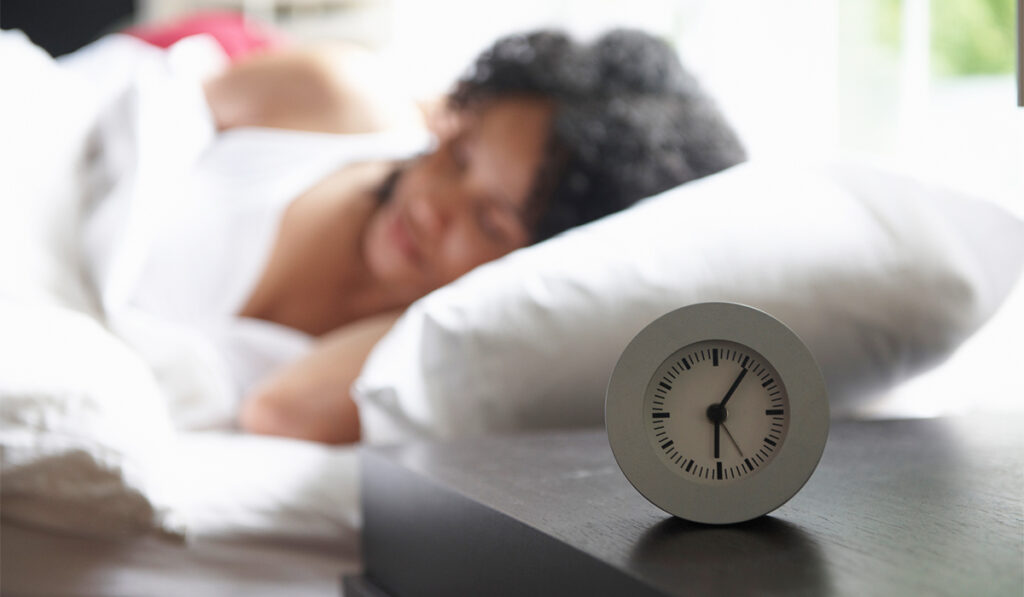 Alarm clock in foreground with soft focus of woman sleeping in bed in background