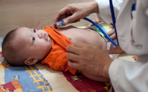 Asian baby getting his heart listened to with stethoscope. Orange shirt. Doctor's white coat.