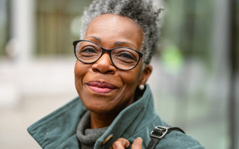 Older smiling black woman with short hair and a green coat, with a purse strap visible over her shoulder and part of her hand. Black glasses. Blurred background.