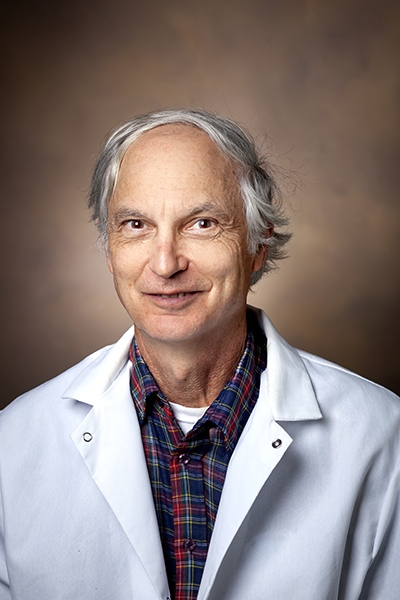 Portrait of Mark Boothby, white coat, plaid shirt, white hair, brown background