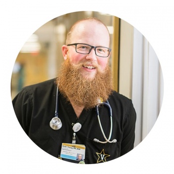 Portrait of Brett Young, in clinical attire, with stethoscope and scrubs, and badge. Big red bushy beard, glasses, bald head.