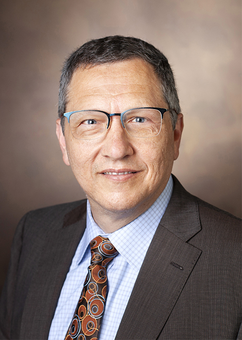 Portrait of Joseph Magliocca, in dark suit, blue shirt and tie. Glasses, dark hair and brown background