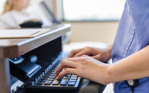 Hands on keyboard in clinical setting, patient out of focus in the background