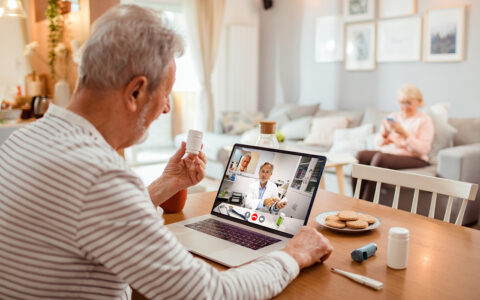 Senior man using telehealth at a dining room table holds up medication bottle to show the doctor