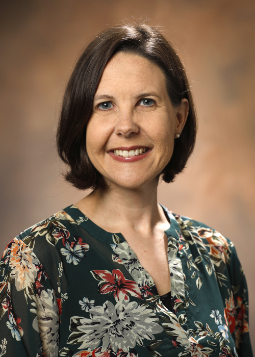Portrait of Amy Hayes, B.S.N. Floral shirt with dark background, chin-length dark hair, smile. Brown background.
