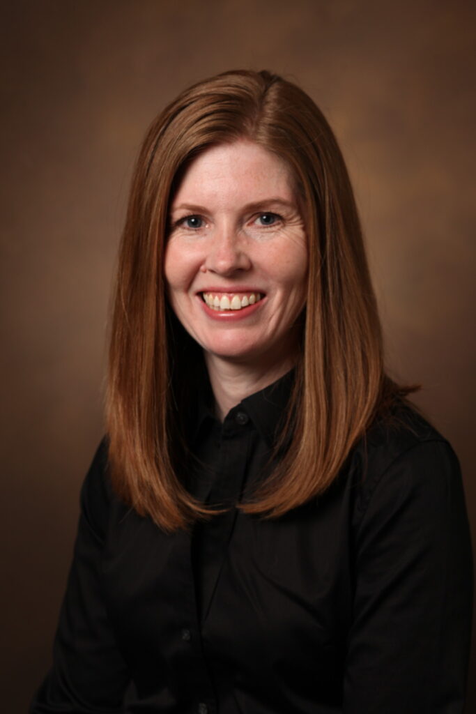 Portrait of Julie Lounds Taylor, Ph.D. Black shirt. Long brown hair, Smile. Brown background.