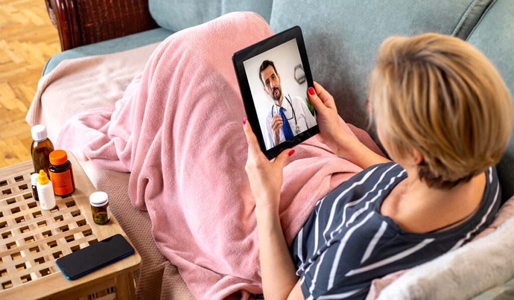 Seated white woman on a couch, wrapped in a blanket, uses telemedicine on a tablet to consult with a doctor