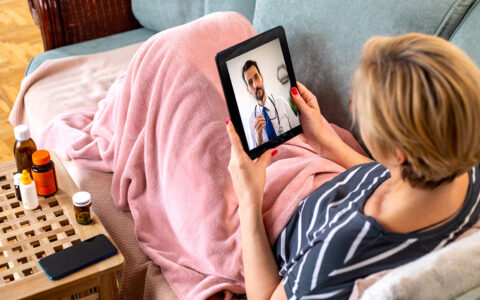Seated white woman on a couch, wrapped in a blanket, uses telemedicine on a tablet to consult with a doctor