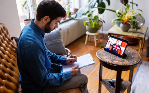 Man sitting on a couch taking notes on a clipboard while videoconferencing with a therapist
