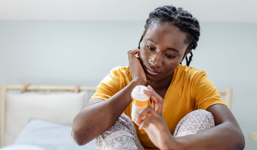 Young African American woman in bedroom looking somberly at prescription pill container