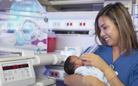 Female nurse feeding newborn Black baby next to an incubator
