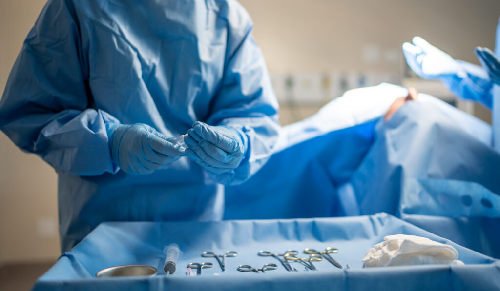 A doctor wearing scrubs and blue gloves chooses an instrument from a surgical instrument table.