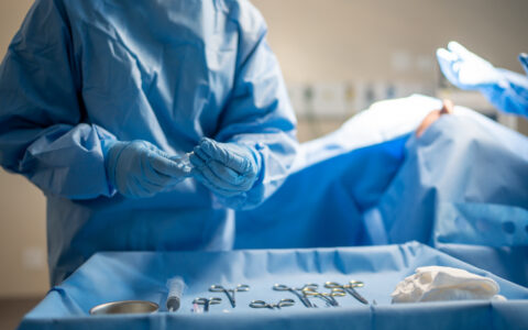 A doctor wearing scrubs and blue gloves chooses an instrument from a surgical instrument table.