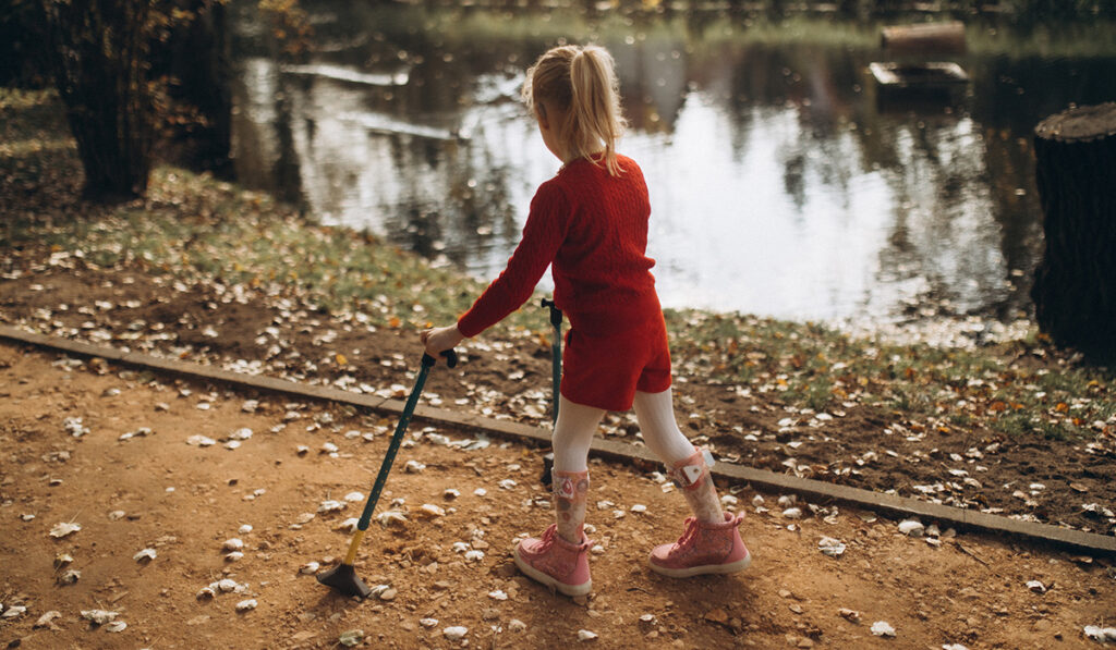 A young girl walks on an outdoor path by a pond, using two canes and leg braces