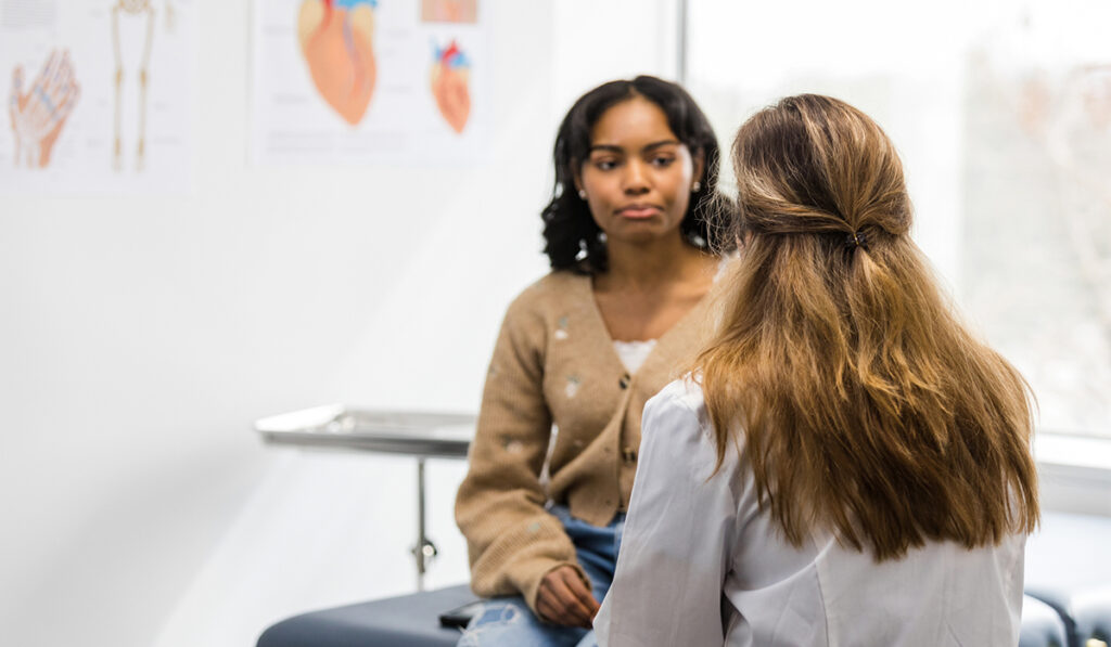 A young woman sits on an examination table at a doctor's office, talking to a female physician.