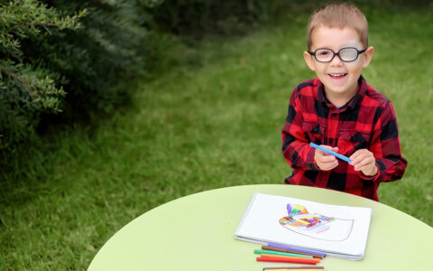 A boy draws a picture seated at at a table in a garden. He wears an eye patch on one eye beneath his glasses to prevent strabismus.