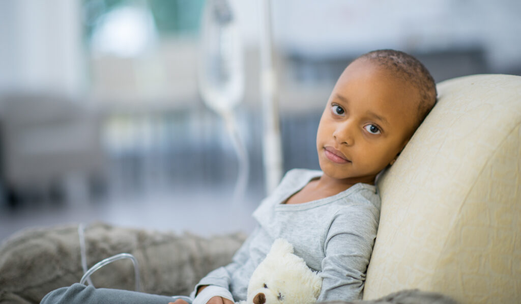 A young girl of African descent is indoors in a hospital room. She has cancer. She is lying in bed with her teddy bear while hooked up to an IV. She is looking at the camera.