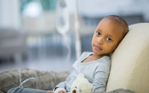 A young girl of African descent is indoors in a hospital room. She has cancer. She is lying in bed with her teddy bear while hooked up to an IV. She is looking at the camera.