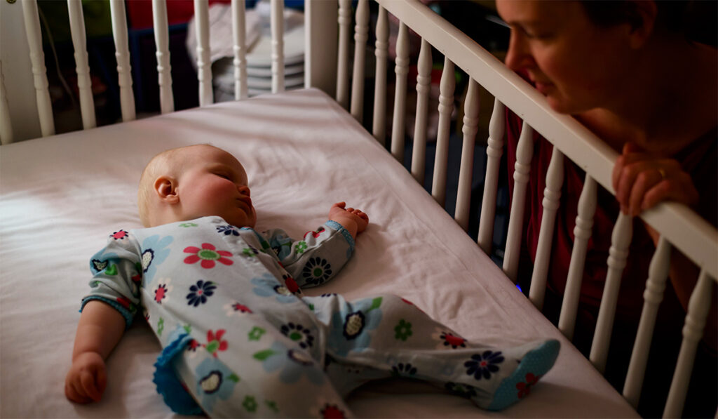 A young woman watches a baby sleeping on her back in a crib