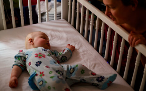 A young woman watches a baby sleeping on her back in a crib