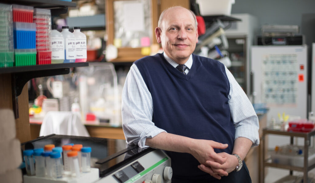 A researcher wearing a sweater vest and tie smiles while leaning against lab equipment.