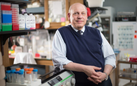 A researcher wearing a sweater vest and tie smiles while leaning against lab equipment.