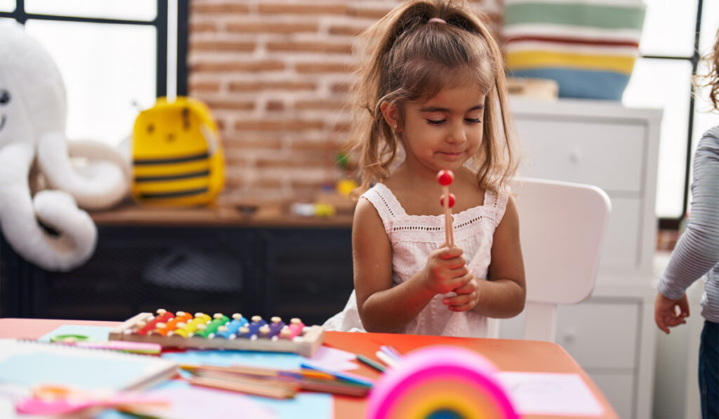 Young Hispanic girl playing xylophone sitting on table at kindergarten