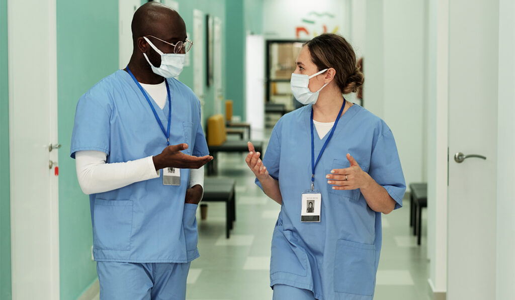 Two young intercultural doctors or surgeons in protective masks having discussion while walking along hospital corridor