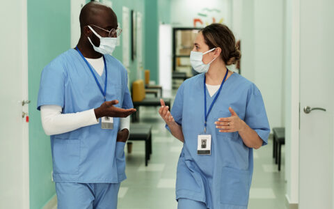 Two young intercultural doctors or surgeons in protective masks having discussion while walking along hospital corridor